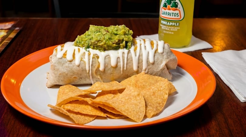 A large burrito topped with guacamole and sour cream, served with tortilla chips and a Jarritos.