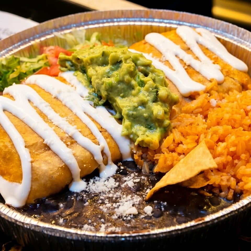 A chimichanga with sour cream, guacamole, rice, and black beans in a foil container.