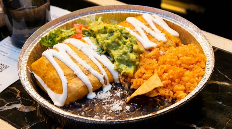 A chimichanga with sour cream, guacamole, rice, and black beans in a foil container.